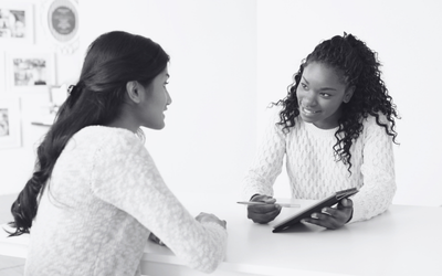 A black and white image of two young women facing each other and talking with each other. One is holding a notebook and gesturing at it with her in pen to the other woman.