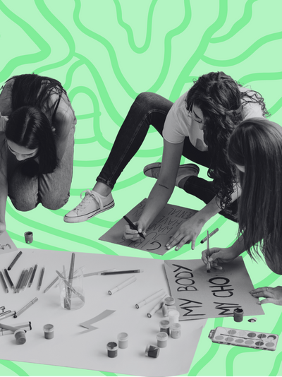A black and white image of three young women sitting on the ground and using markers and paints to create protest signs.