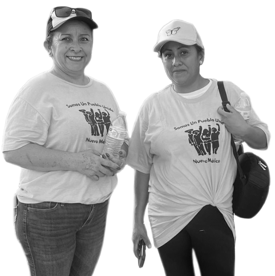 A black and white image of two middle-age medium skin women wearing matching Somos Un Pueblo Unido t-shirts, standing and smiling.