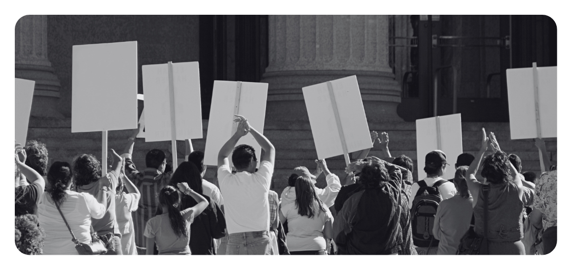 A black and white image of the backs of a crowd of protesters, holding signs, with some holding their hands up in the air and clapping.