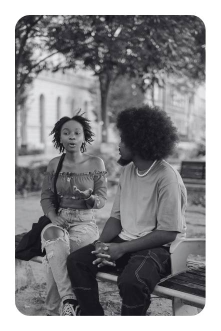 A black and white image of a young, dark skin woman sitting on a bench and talking, gesturing with her hand, while a young, dark skin man sits next to her on the bench, listening.