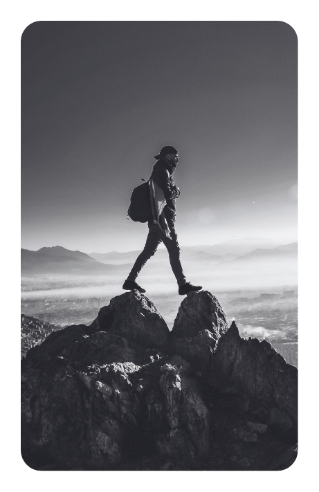 A black and white image of a young light skin man straddling two rocks on top of a mountain, with the views of a mountain range behind him.