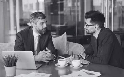 A black and white image of two men in suits sitting at a table talking with each other. One man has a laptop open in front of him, while the other is writing in a notebook. They both have coffee cups in front of them.